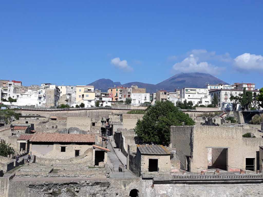 Figura 1 - In primo piano, la città romana di Herculaneum (Ercolano), distrutta dall’eruzione del 79 d.C. del Vesuvio, visibile sullo sfondo. Fra la città romana e il vulcano, le case della moderna Ercolano. Foto di Micol Todesco.